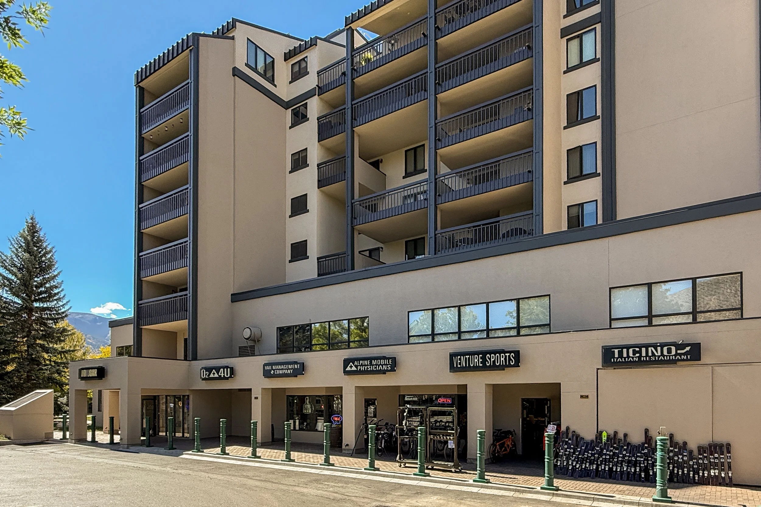 A multi-story building with commercial spaces on the ground floor and residential units above. The ground floor includes signs for businesses such as an Italian restaurant, a sports shop, a mobile company, and other shops. The building has balconies 