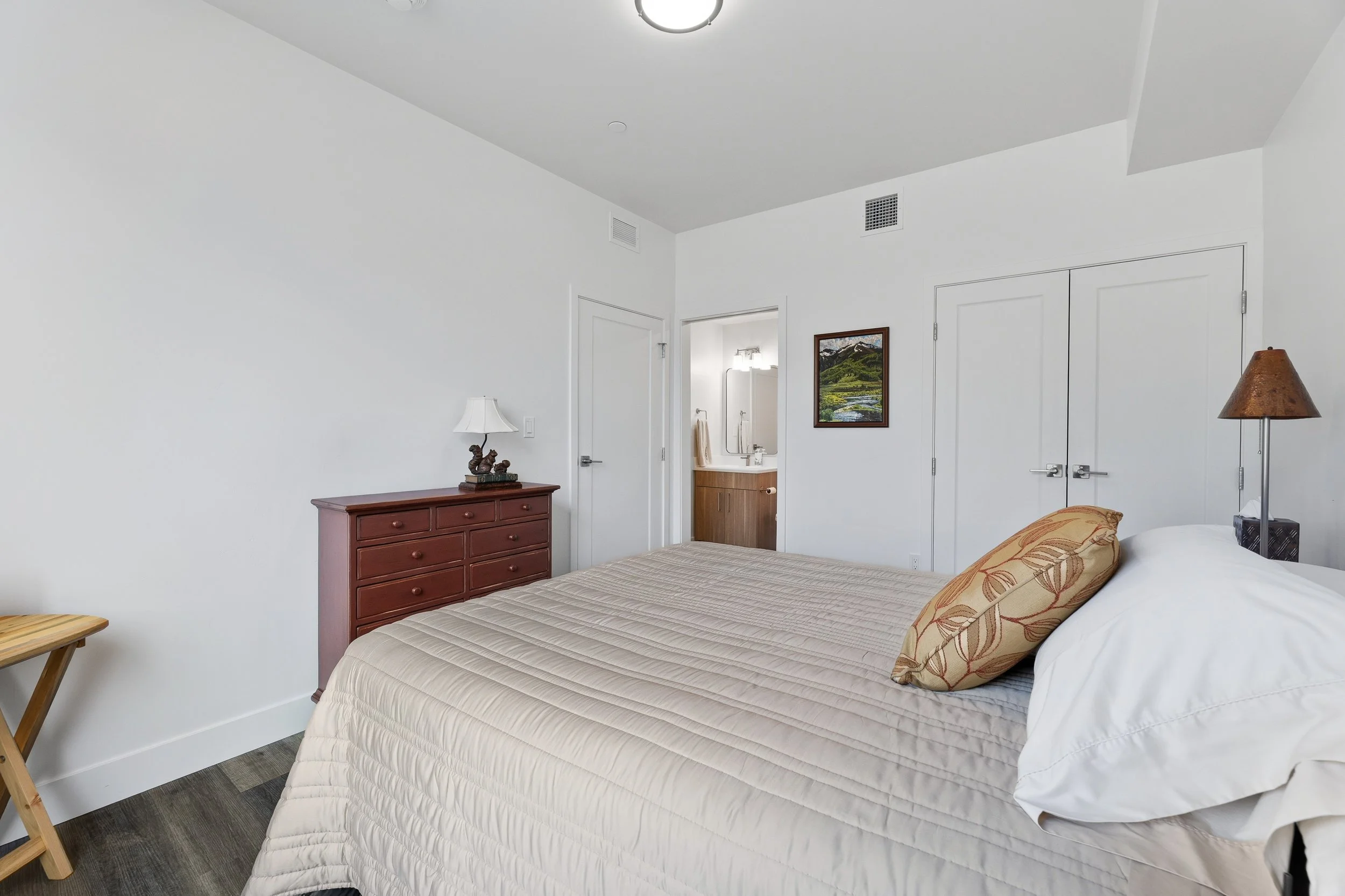 A bedroom with white walls, a bed with beige bedspread and floral pillows, a wooden dresser with a lamp, a small wooden folding table, a framed landscape painting, a closet with double doors, and a bathroom visible through an open door, all with mode