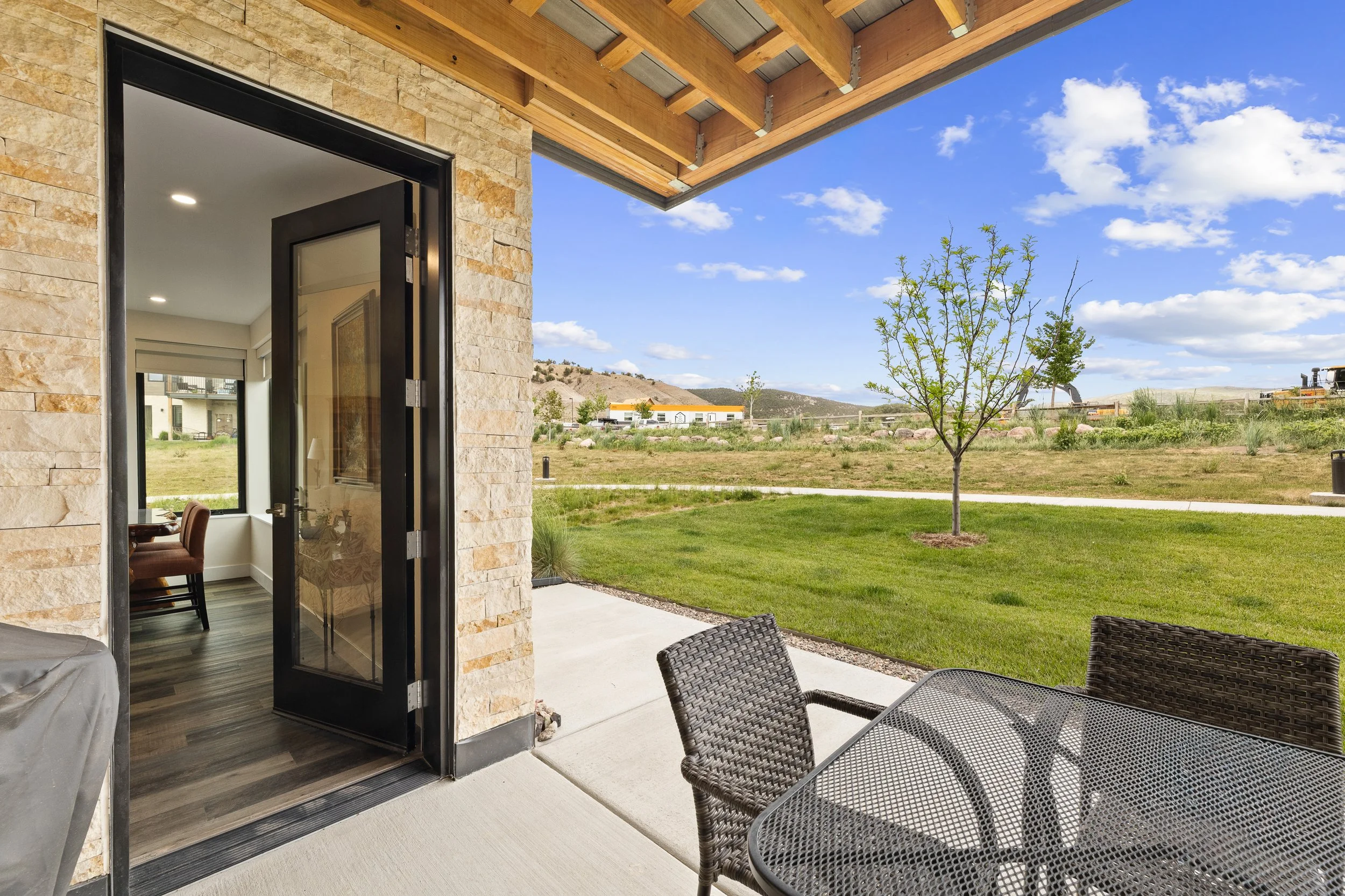 A patio with outdoor furniture including a glass-top table and woven chairs, overlooking a green lawn with small trees and hills in the background under a blue sky with clouds. The patio is attached to a house with stone exterior walls and an open do