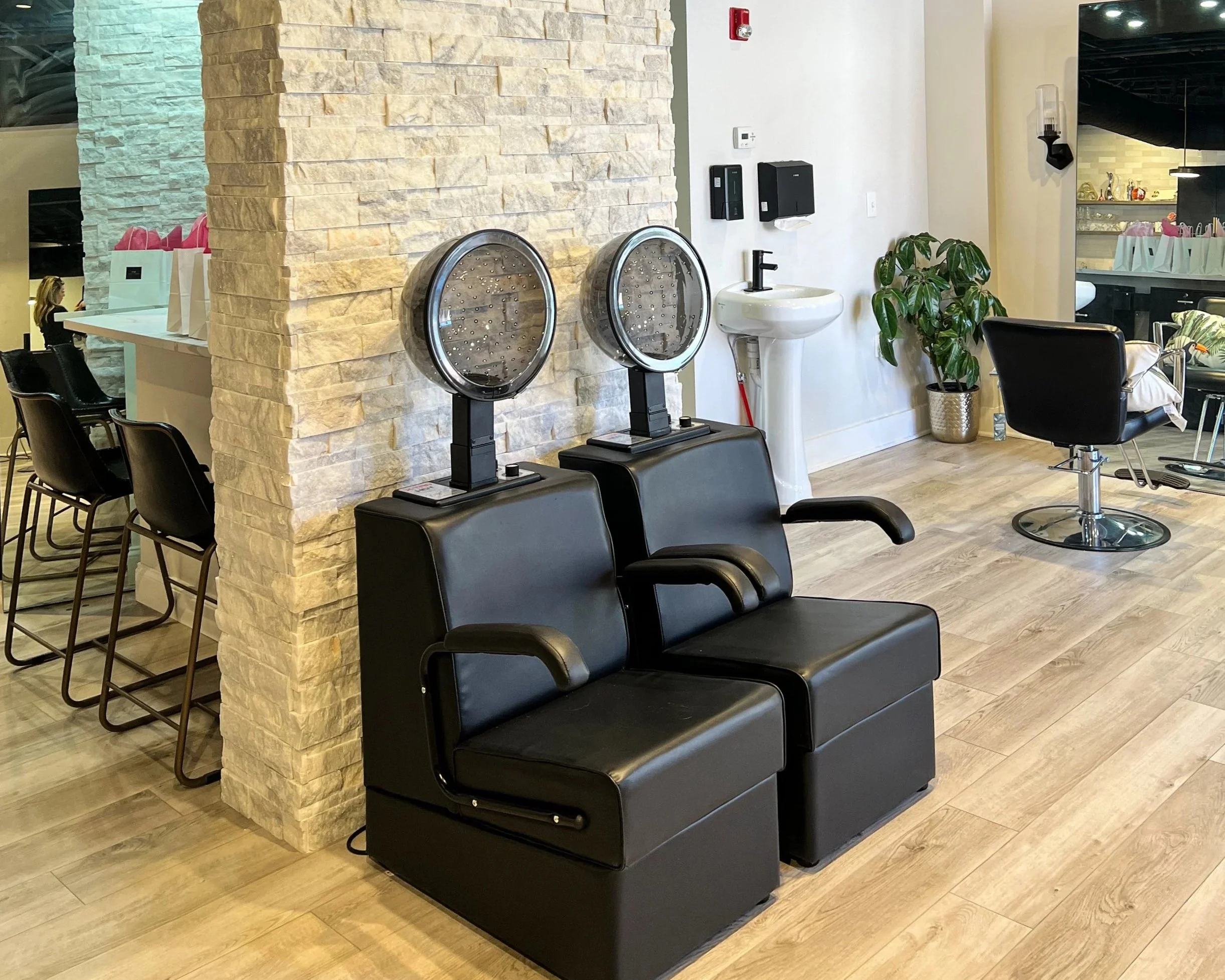 Hair salon with two black hair drying chairs with hoods and a barber chair, a small sink, and a potted plant.
