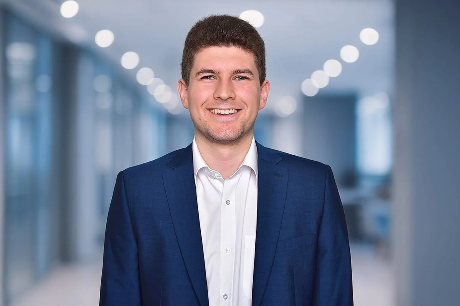 A smiling man wearing a navy blue suit and white collared shirt, standing in a bright, modern hallway with large windows and circular ceiling lights.