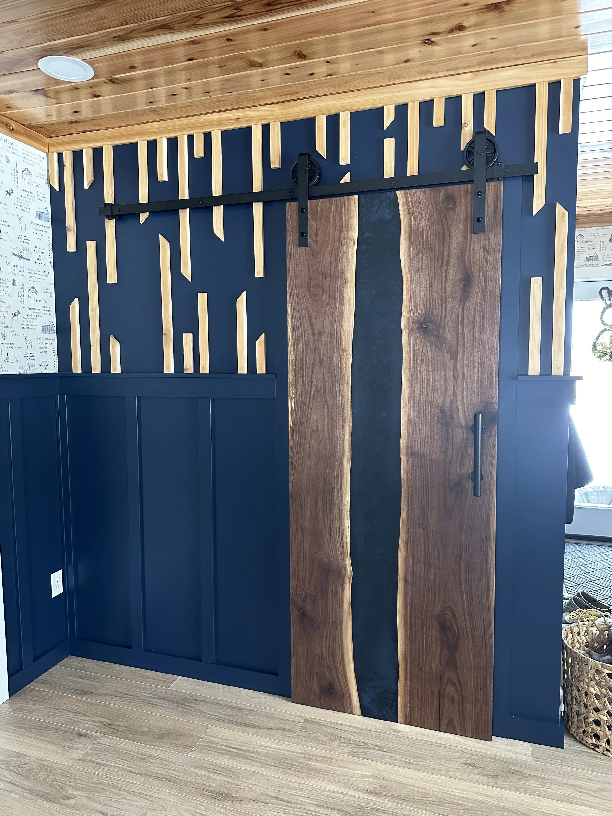 Interior with a sliding barn door featuring a live edge wood design, set against a navy blue wall with vertical wooden accents. The ceiling and floor are made of a lighter wood, creating a rustic aesthetic.