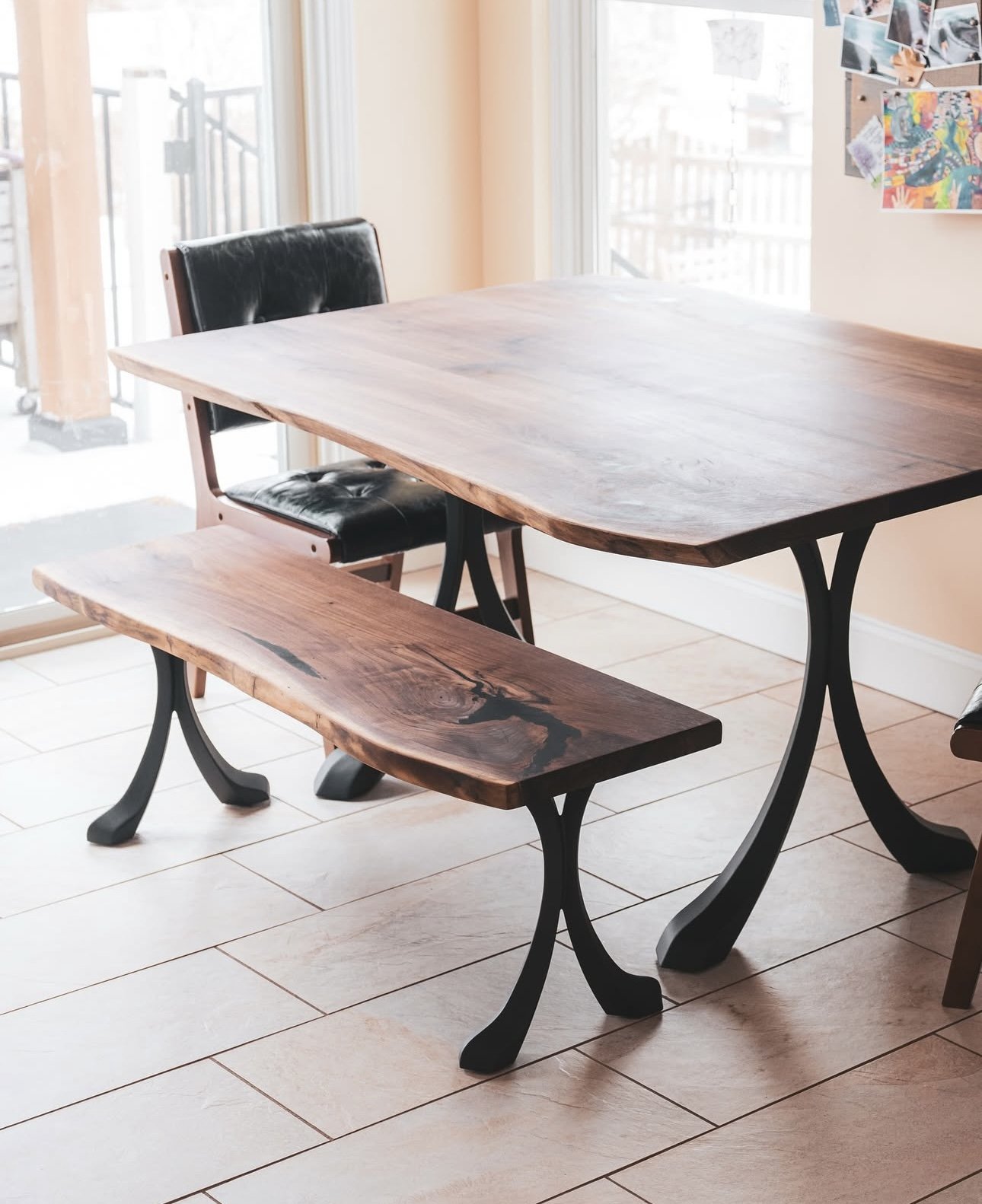Wooden dining table with a matching bench and a chair in a sunlit room with tiled flooring.