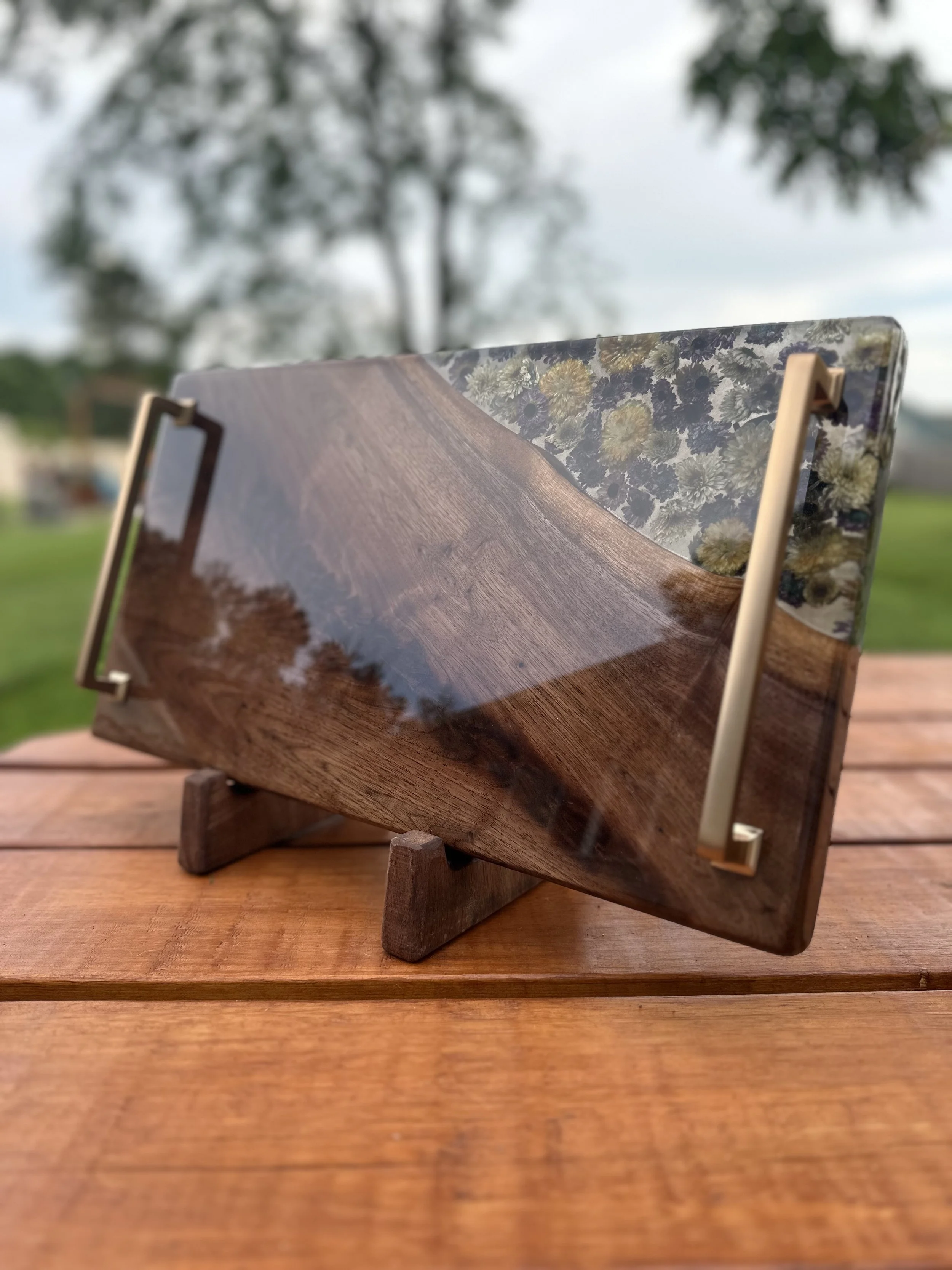 Wood and resin serving tray with metal handles on a wooden stand, featuring embedded flowers and a natural finish, outdoor setting.