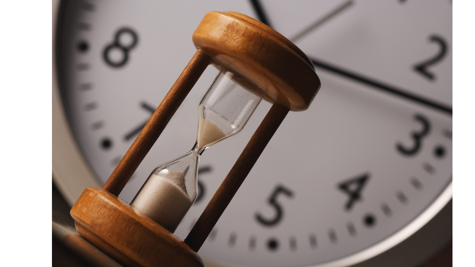 Close-up of an hourglass with white sand, positioned in front of a blurred round wall clock showing partial numbers from 8 to 2.