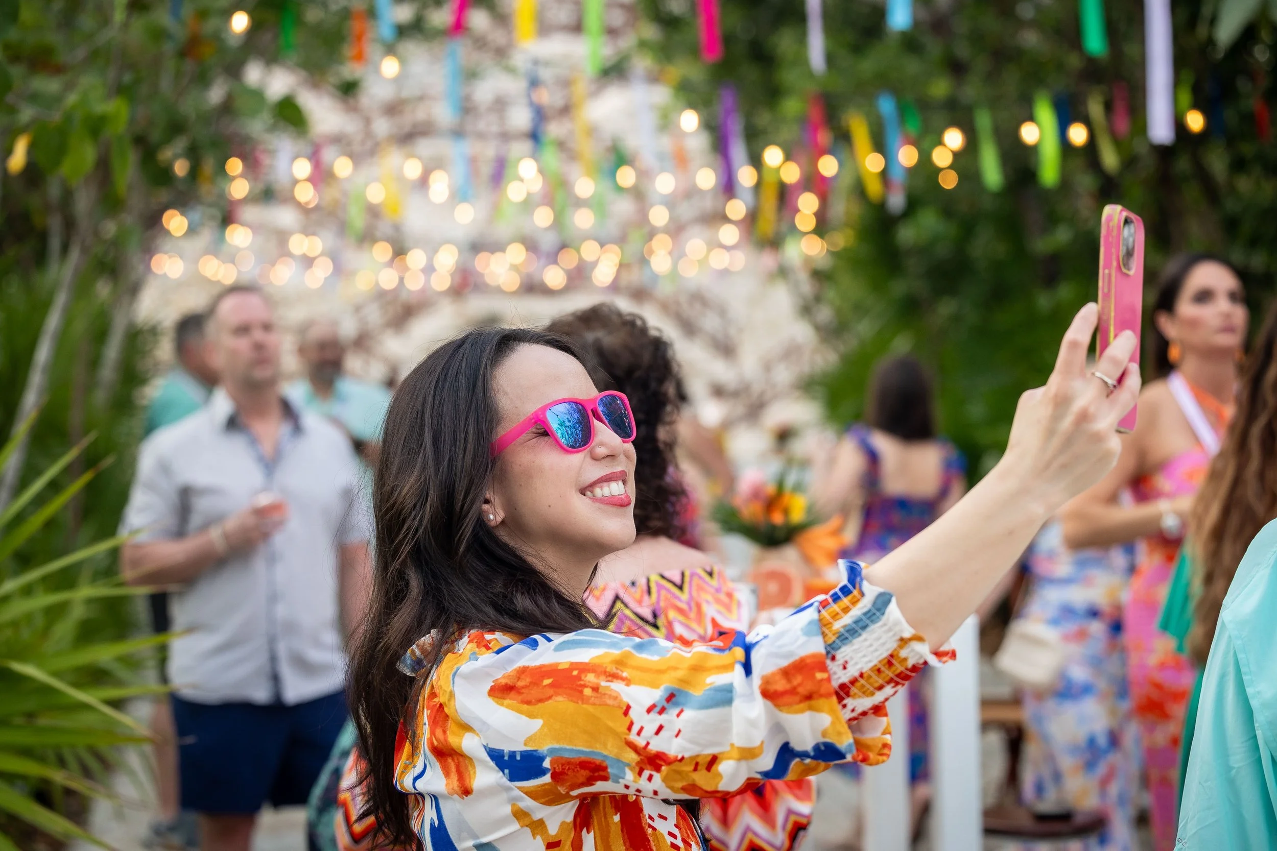 Woman wearing pink sunglasses smiling and taking a photo with a pink phone at an outdoor party with colorful decorations.