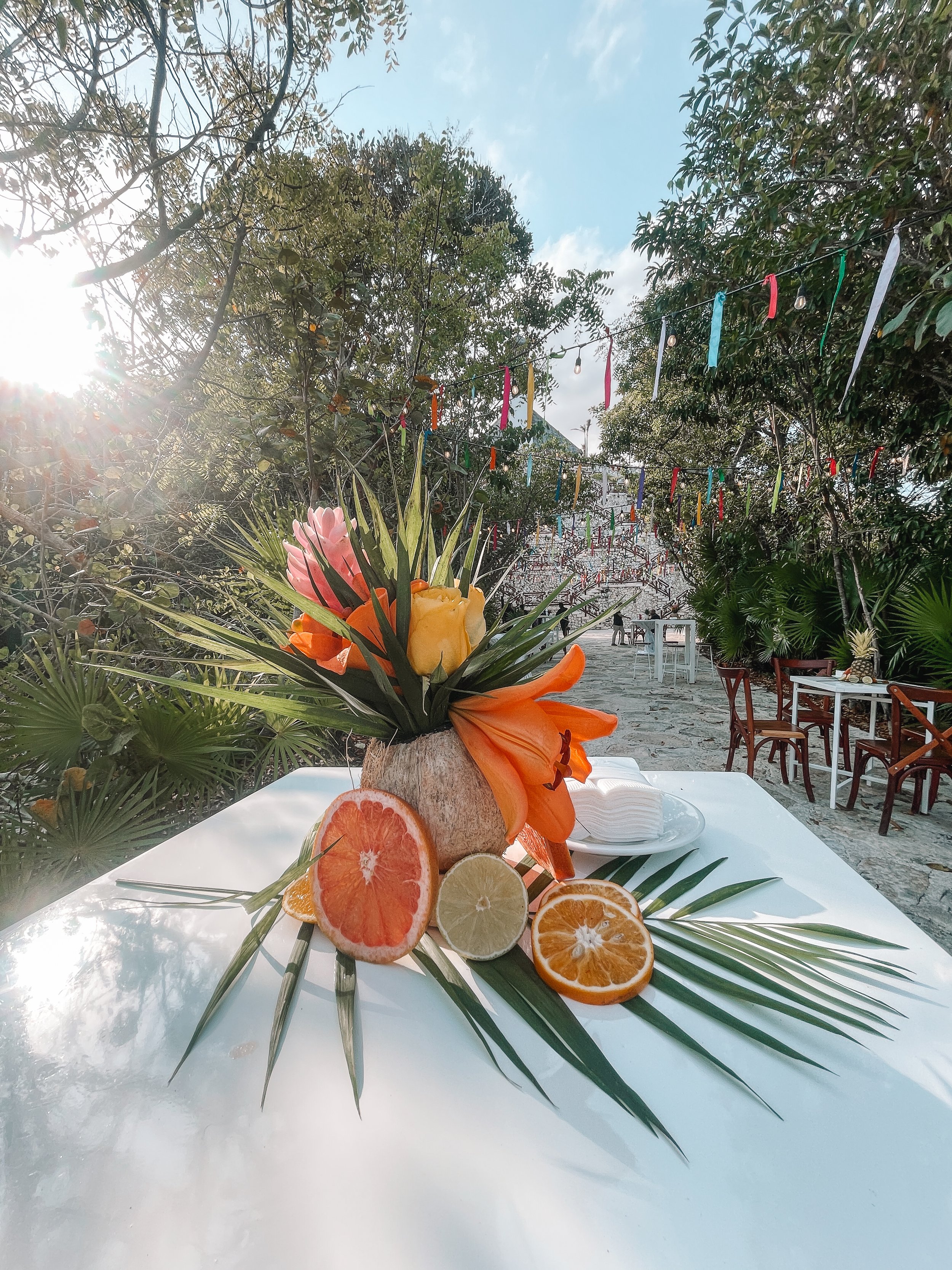 A table with a floral centerpiece made of tropical flowers and citrus fruits, set outdoors with trees and colorful decorations hanging above for a celebration reception.