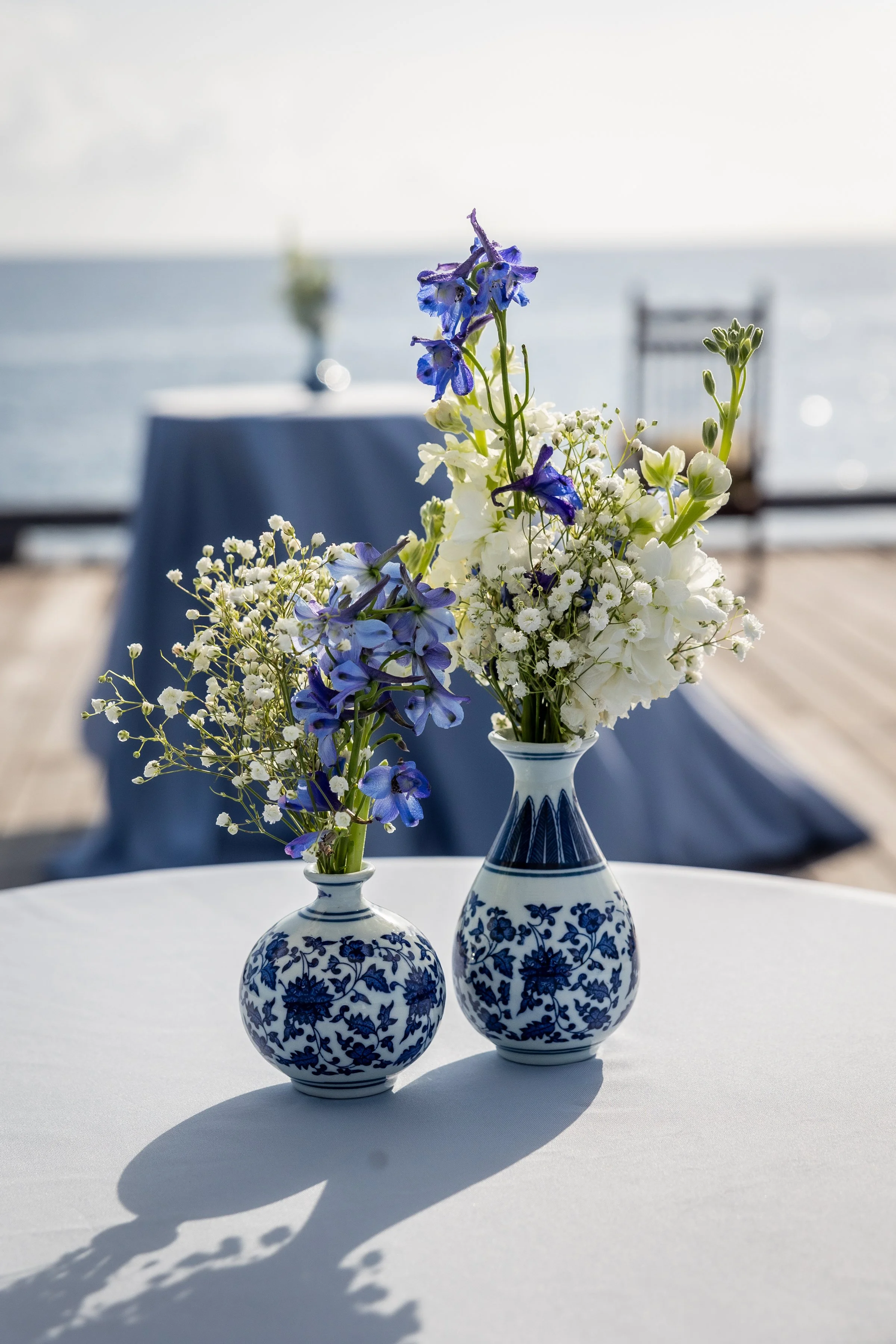 Two blue and white floral vases with flowers on a white table, overlooking a body of water with a blurred table and chair in the background for a corporate incentive event in Nives.