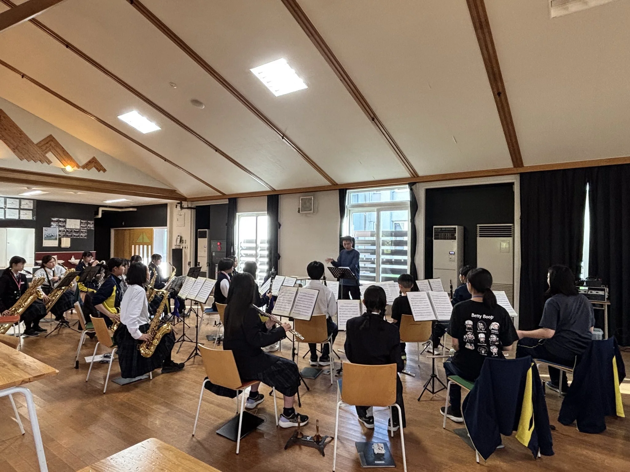 A group of children in a music classroom playing saxophones, guided by a conductor. The classroom has wooden floors, large windows, and posters on the walls.