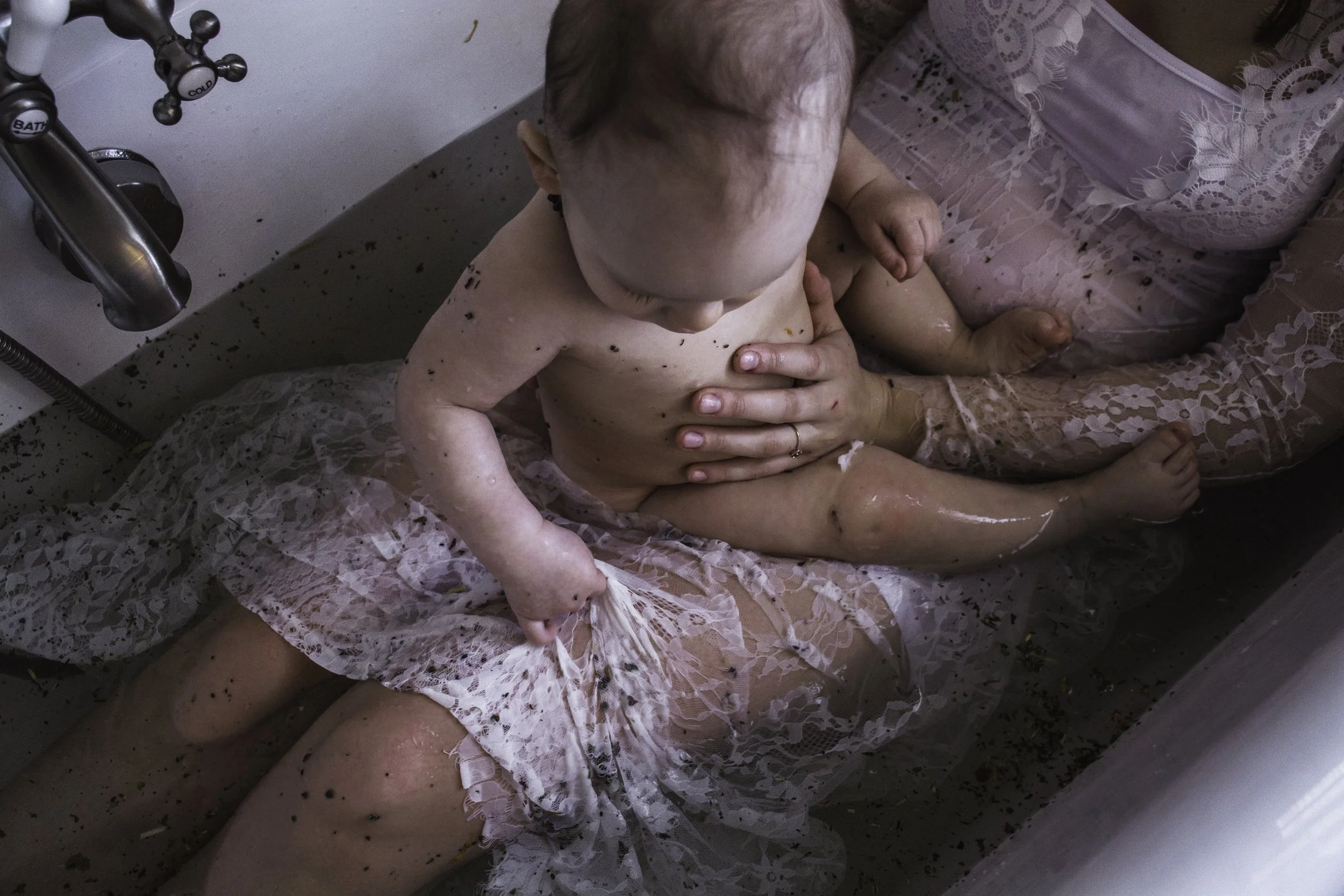 A baby sitting in a small bath tub during bath time, with a woman holding the baby from behind. The baby is surrounded by water and soap suds, and the woman is dressed in a pink lace dress.
