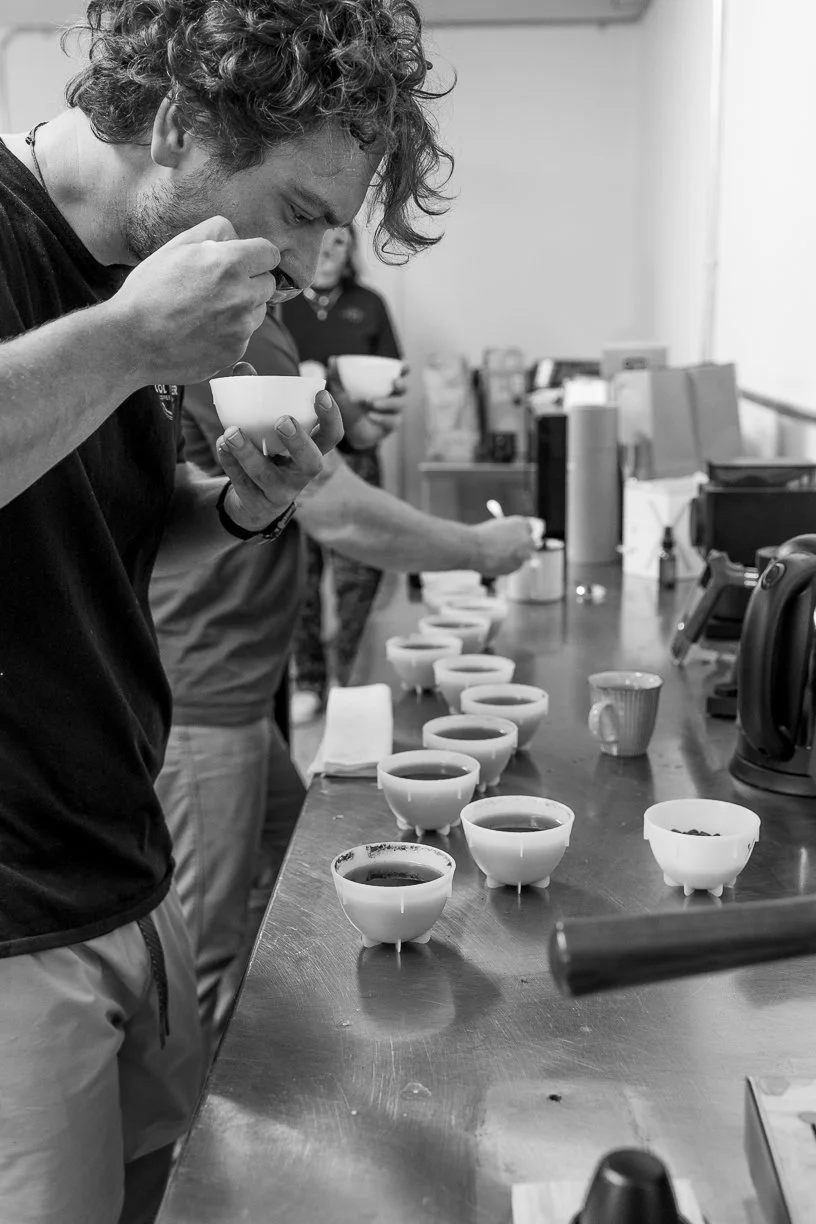 A man tasting coffee in a row of coffee cups on a counter, with another person in the background also holding a cup.