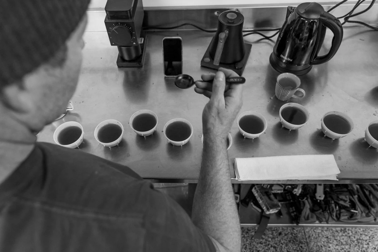 A person is conducting a coffee tasting, with multiple cups of coffee in front of him on a table, using a spoon to sample the coffee. Various coffee brewing equipment and a cup with a hot beverage are visible on the table.