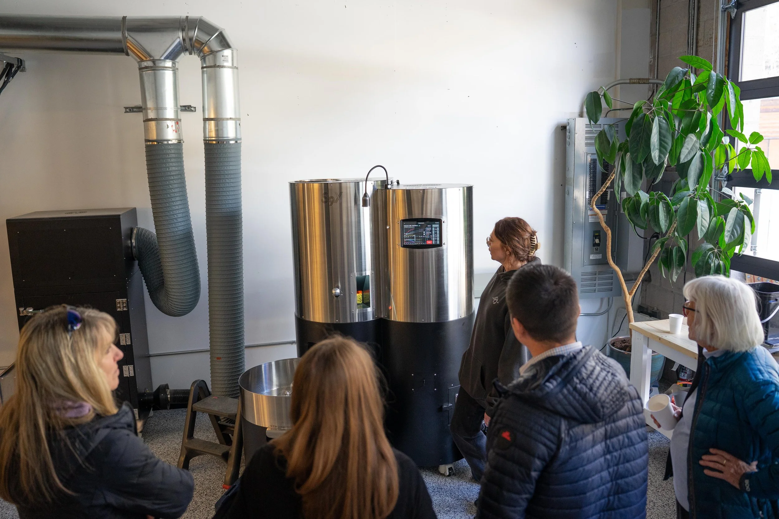 A group of people listening to a woman explaining a coffee roasting machine inside a modern industrial-style space with a large window and a potted plant.