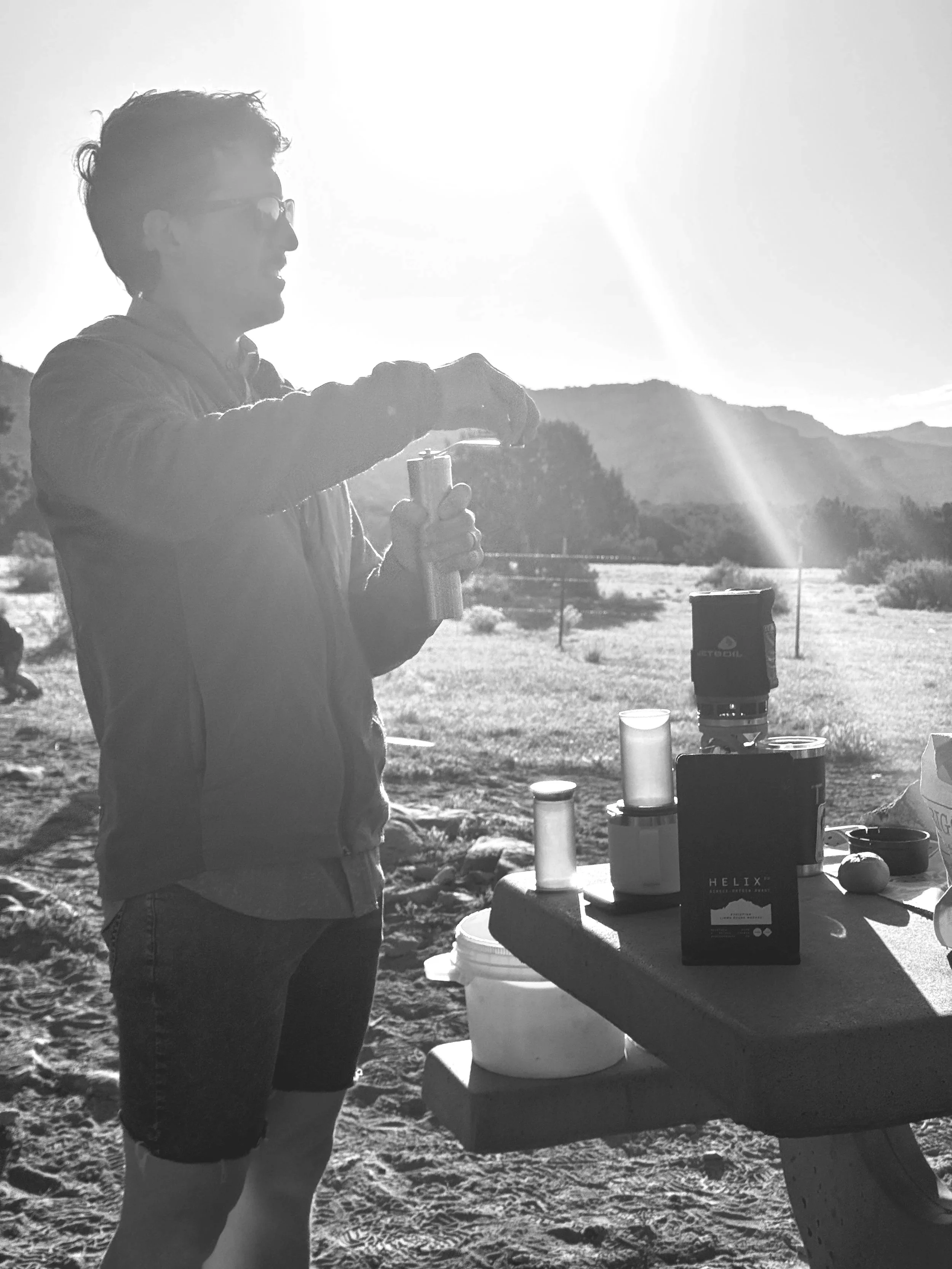 A man standing outdoors in bright sunlight, preparing a drink or snack at a picnic table. The landscape features mountains and open fields in the background.