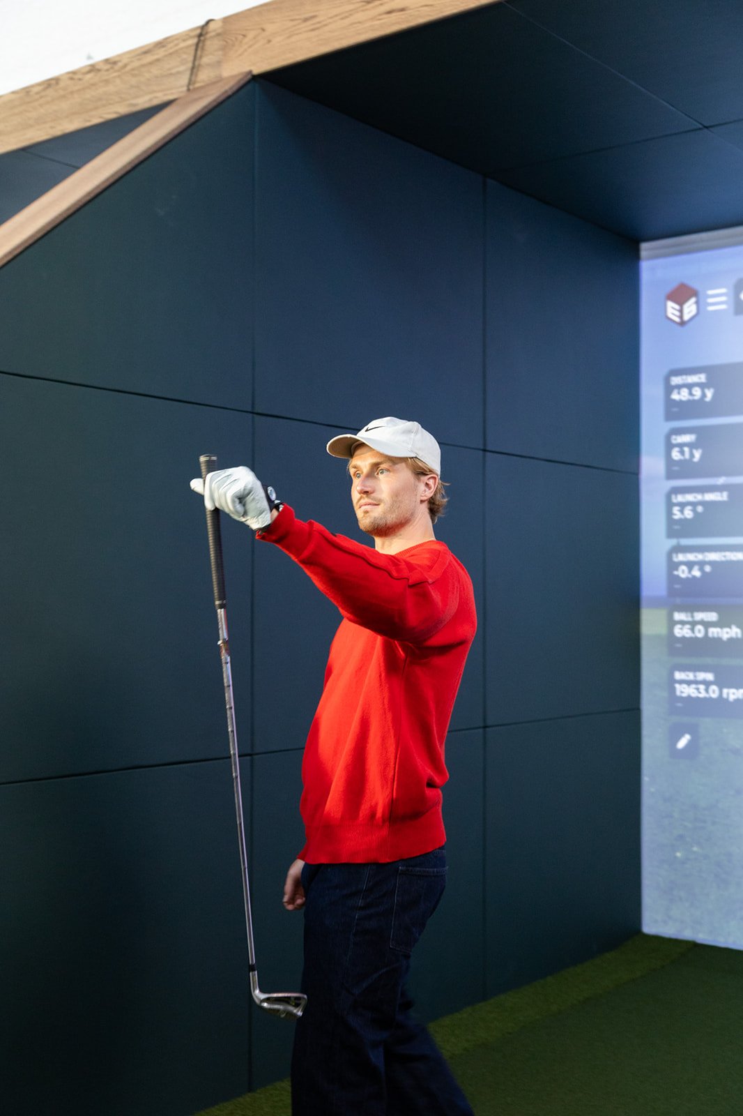 A man wearing a red sweater and a white cap is testing a golf ball in a golf simulator with a screen that displays data.