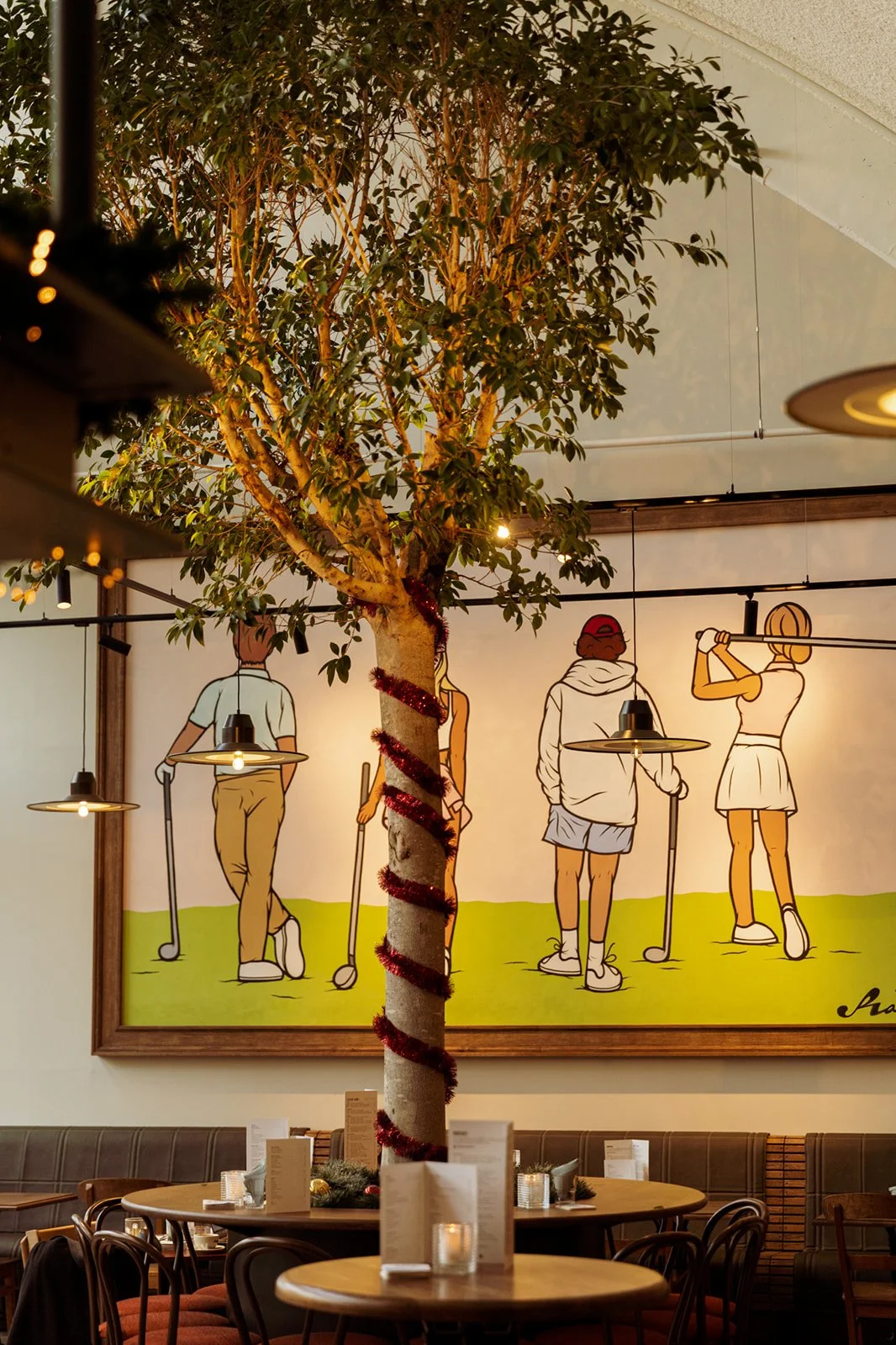 The interior of a restaurant featuring a piece of art depicting four cartoon characters playing golf, an artificial tree with lights, and Christmas decorations on the trunk.