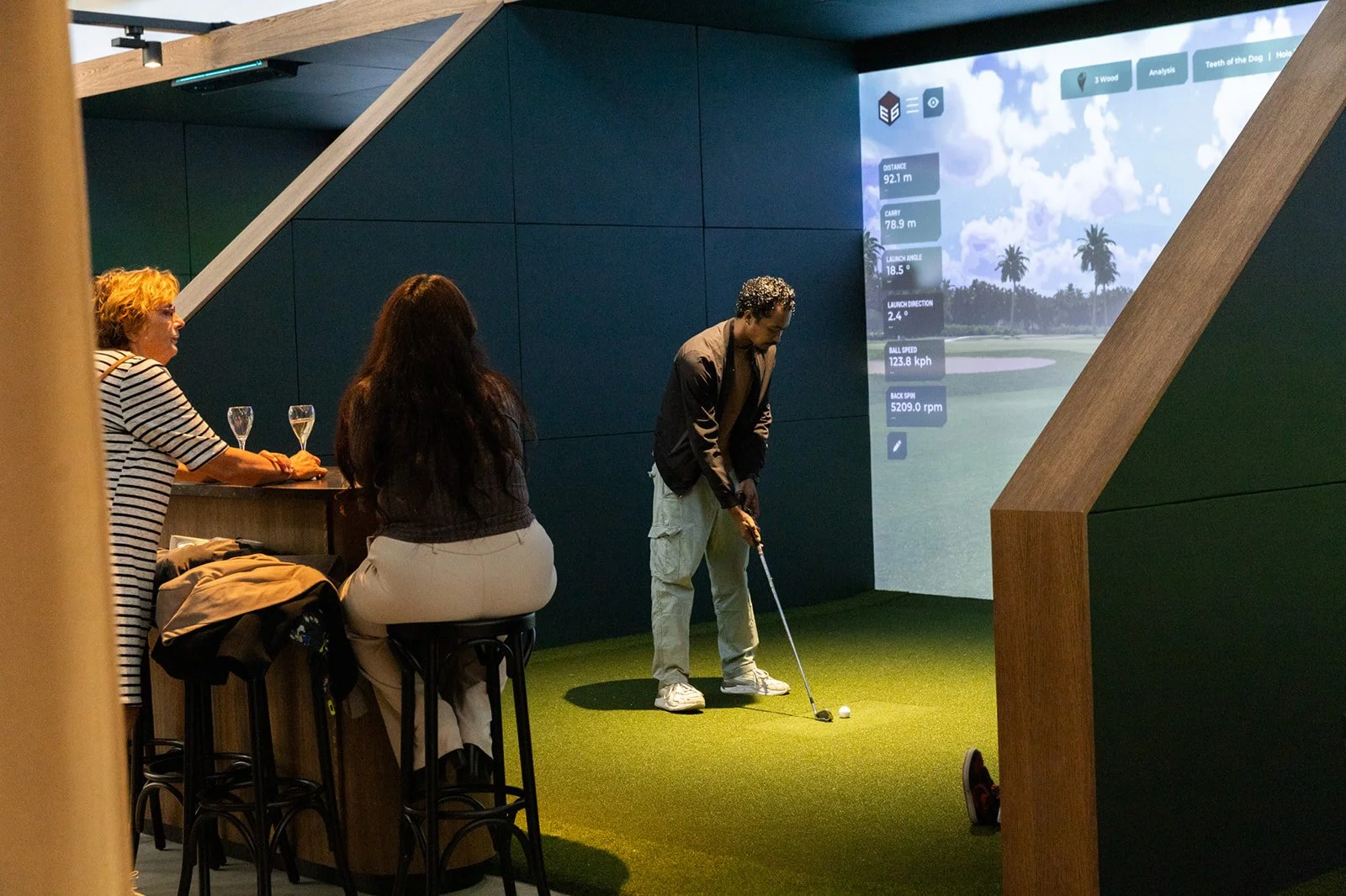 An indoor space with a virtual golf simulator; three people are watching as a man hits a golf ball on artificial turf; two women are sitting, one of whom is holding a glass of wine.