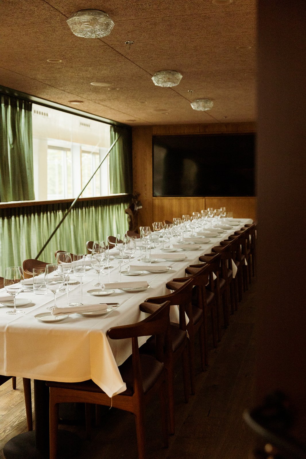 A long table with a white tablecloth, cutlery, wine glasses, and plates, set for dinner, in a room with wooden walls and green curtains.