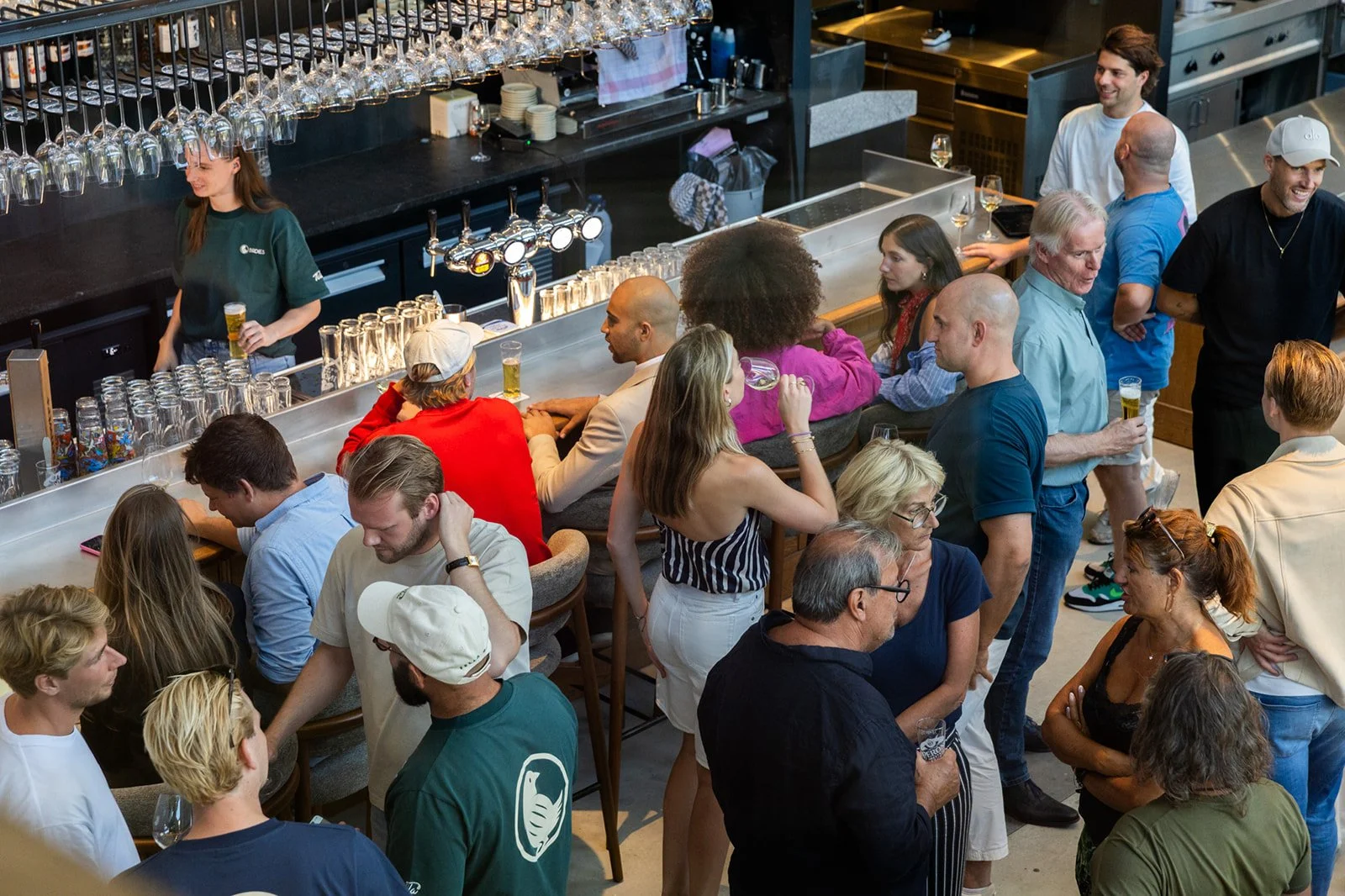 People standing at a long table in a bar, having a drink, while bartenders stand behind the bar, in a bustling and lively setting.