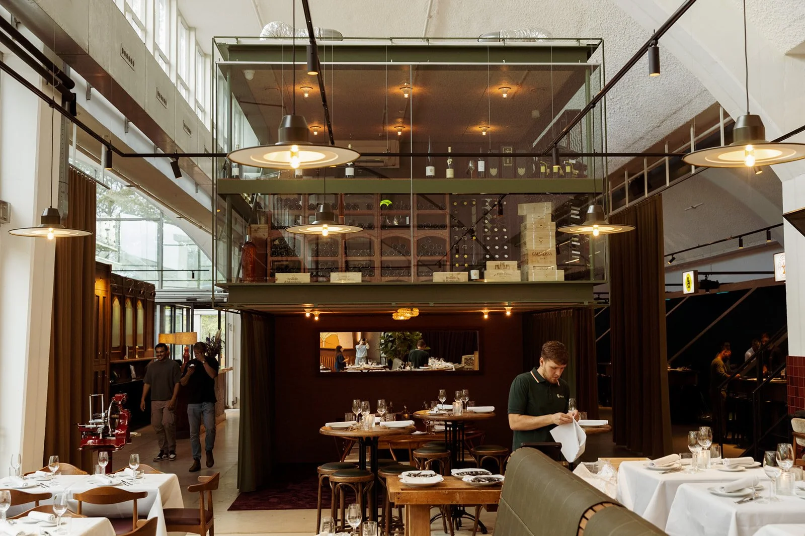 Interieur van een modern restaurant met gedekte tafels, een ober die serviette uitpakt, en een loungebar op de achtergrond met wijnrekken en kookspullen.