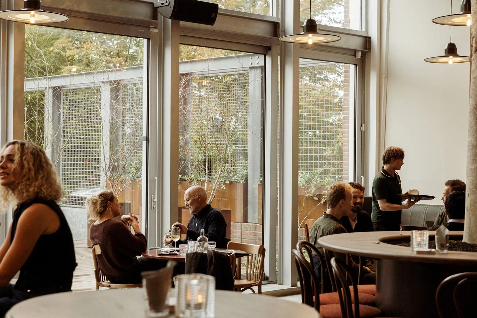The interior of a modern restaurant with large windows, where people are sitting, dining, and chatting while a waitress brings out food.