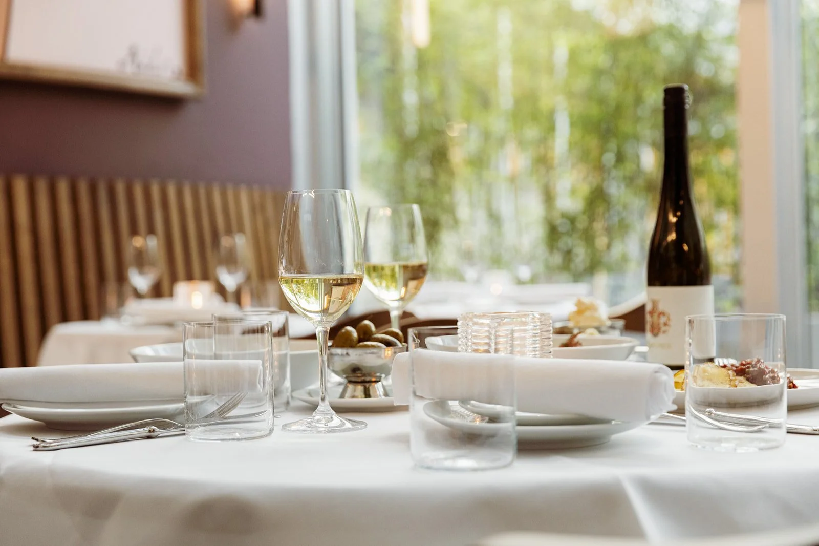 An elegant dining table set with a white tablecloth, glasses of white wine, plates, cutlery, and a bottle of wine, ready for dinner. A green garden is visible through the window.