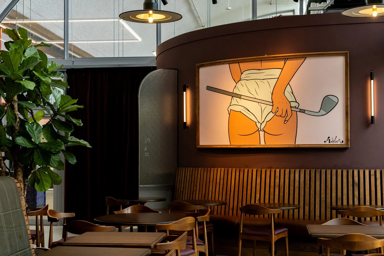 The interior of a modern restaurant featuring wooden tables and chairs, a large plant, and a piece of art on the wall depicting a person holding a golf club and preparing to take a swing.