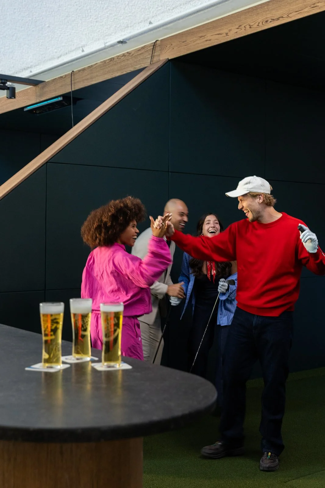 Four friends are laughing and high-fiving each other while playing golf at an indoor sports facility, with four glasses of beer on the table.