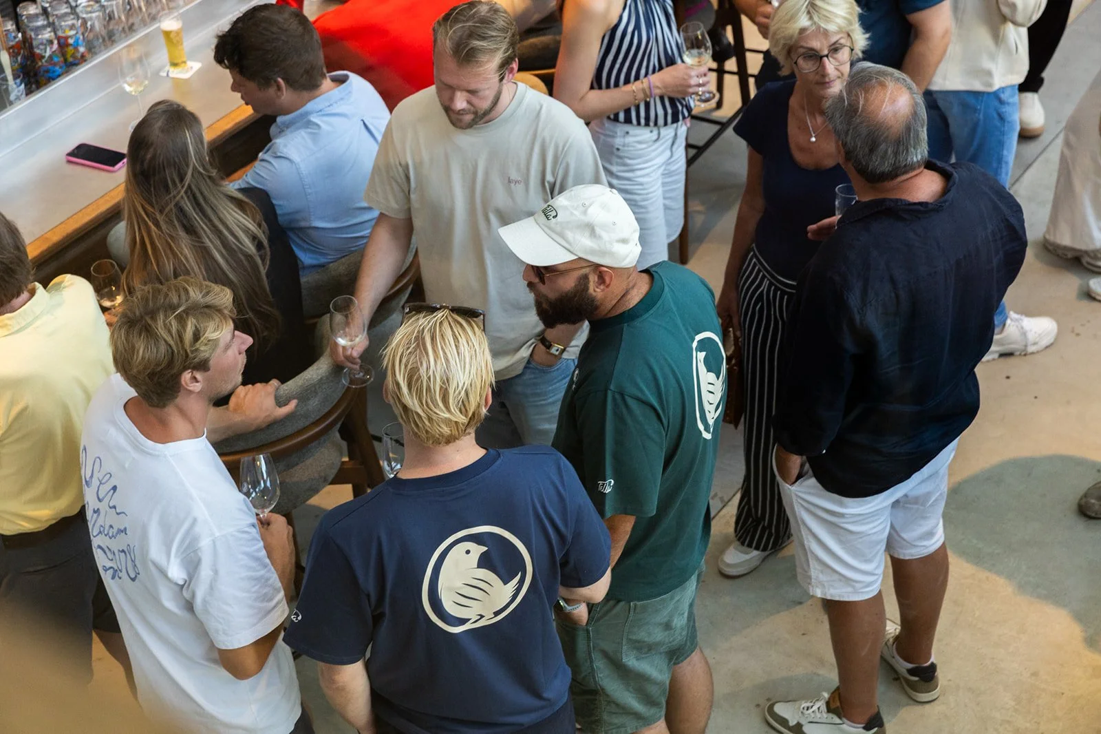 A group of people at a lively social gathering, with beer, wine, shot glasses, a bar, casual attire, and a group engaged in conversation.
