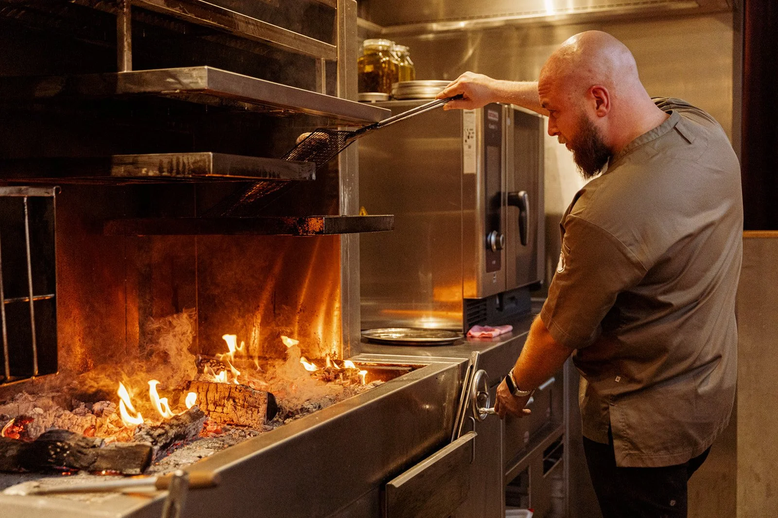 A man in a restaurant kitchen is tending an open-fire grill.
