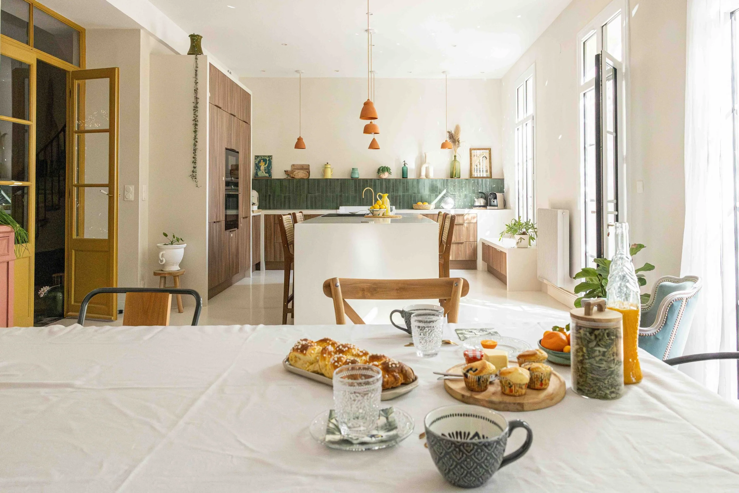 Intérieur lumineux d'une cuisine moderne avec une table en mise en place pour le petit-déjeuner, comprenant des verres, une brioche, des muffins, du jus d'orange et des plantes.