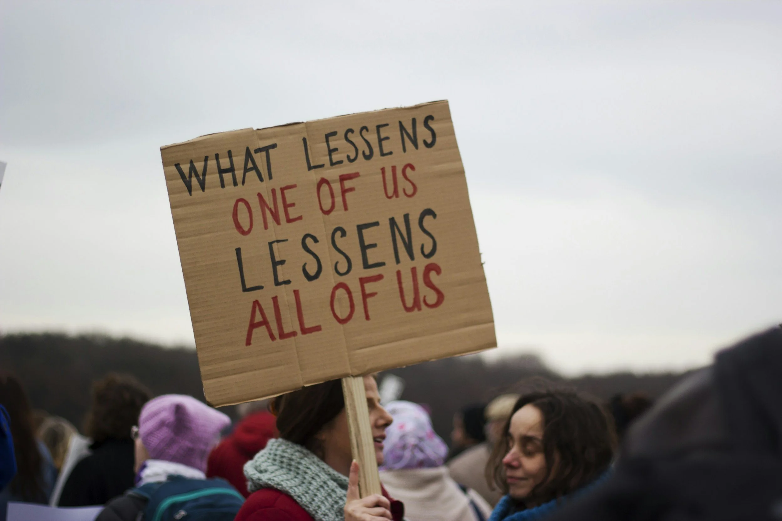 A woman holding a cardboard sign that reads 'What lessons one of us learns all of us' during a crowd gathering, with various people surrounding her, some wearing hats and scarves, under an overcast sky.
