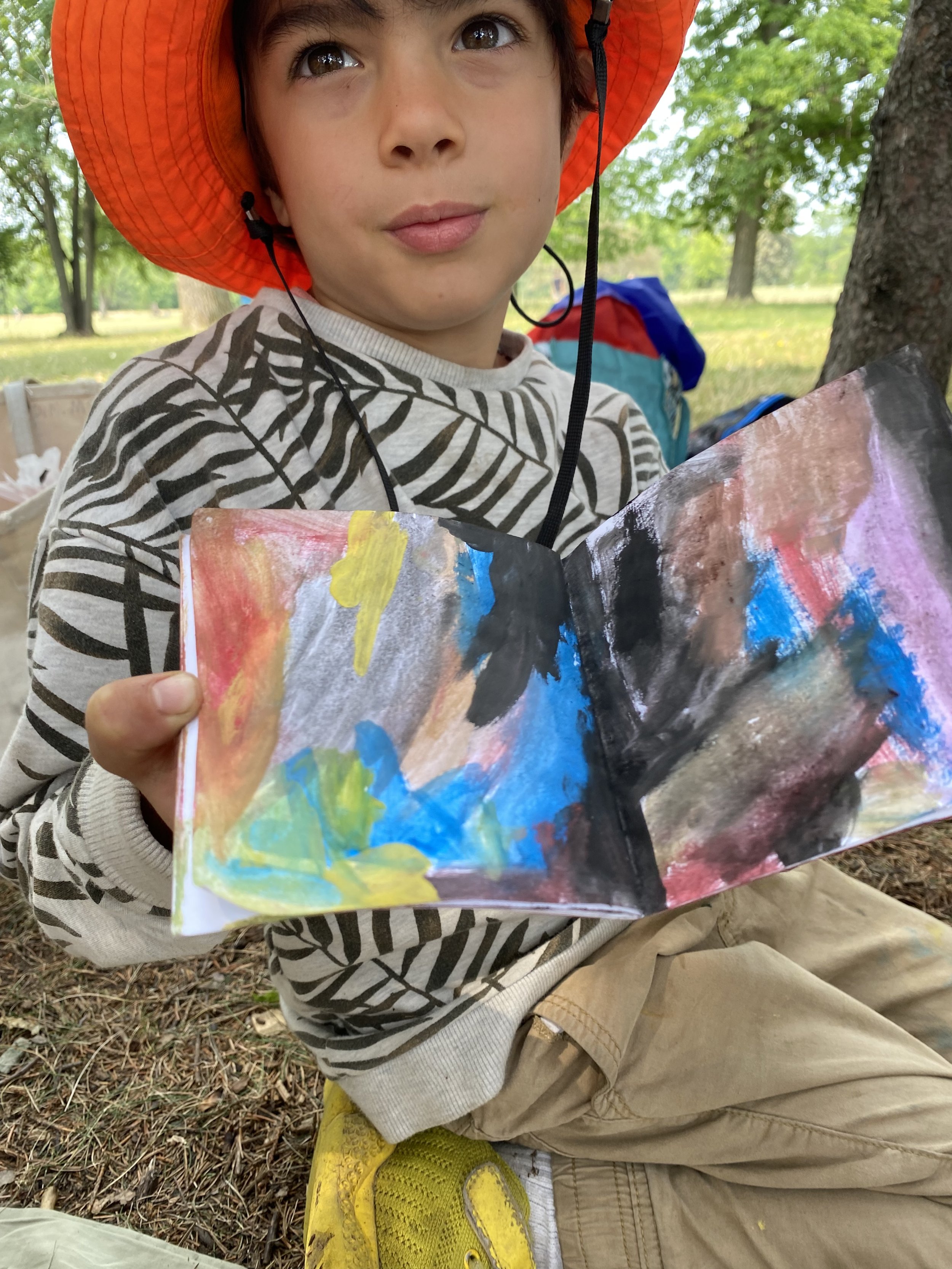 Child wearing an orange hat showing a colorful abstract painting outdoors
