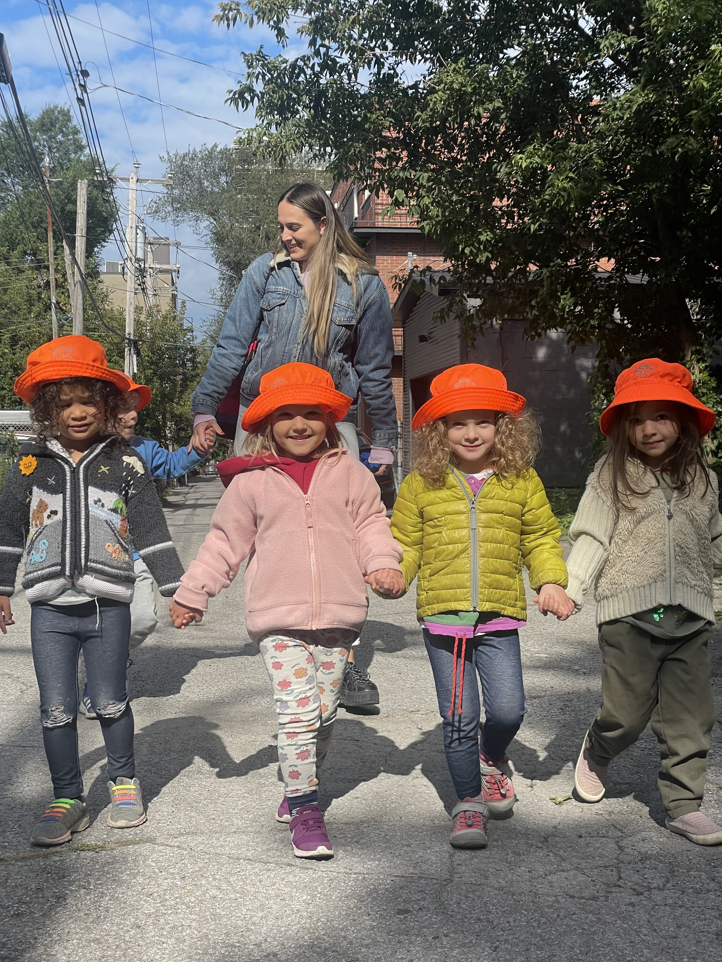 Group of children wearing orange hats and colorful jackets walking together on a street with an adult woman.