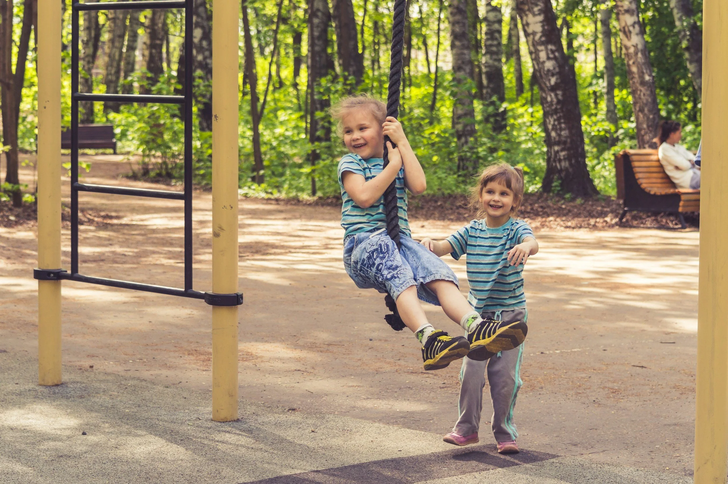 Two children playing on a rope swing at a playground in a park, with trees in the background.