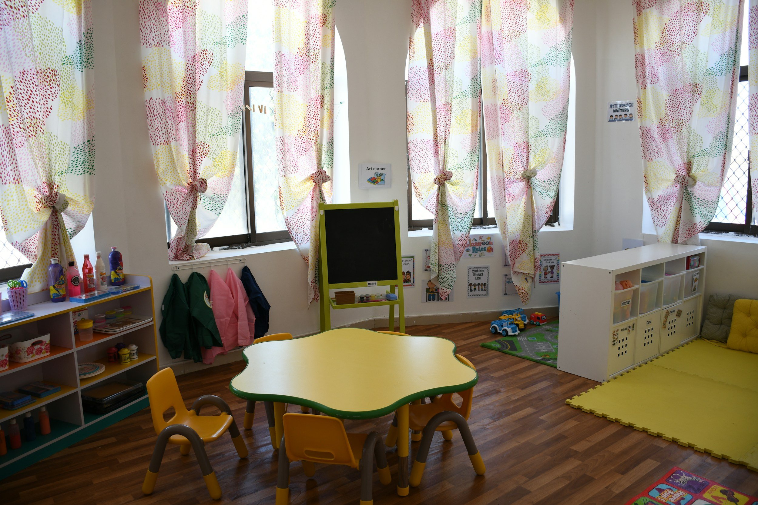 Bright and colorful classroom for young children with large windows and patterned curtains, a yellow and green table with matching chairs, a blackboard on an easel, toy shelves, and storage bins. The floor is wooden with a yellow play mat and a small