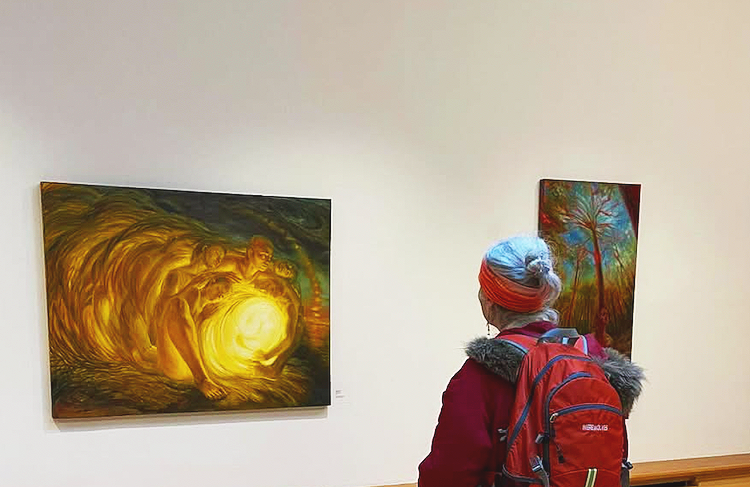 A woman with a red backpack and a head covering looking at colorful paintings in an art gallery.
