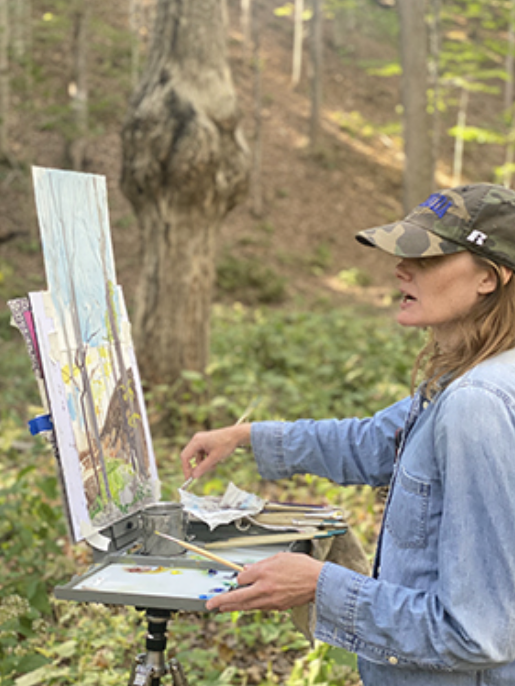 A woman painting outdoors in a forest. She is wearing a camouflage cap and a denim jacket, and is using a portable easel with a picture of trees and a sky.