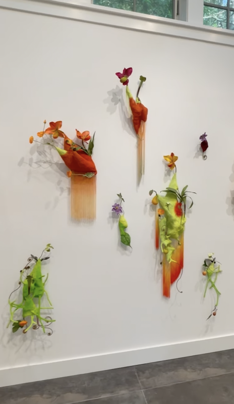 Colorful flower art installation on a white wall featuring paper mache flowers in various colors and sizes attached to wooden blocks and strands.