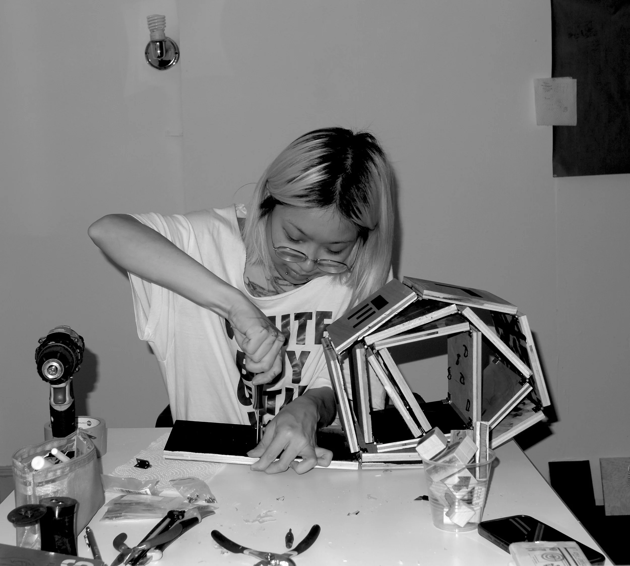 A person with blonde hair and glasses working on a small robotic project at a cluttered table, surrounded by tools and parts, with a plain wall in the background.