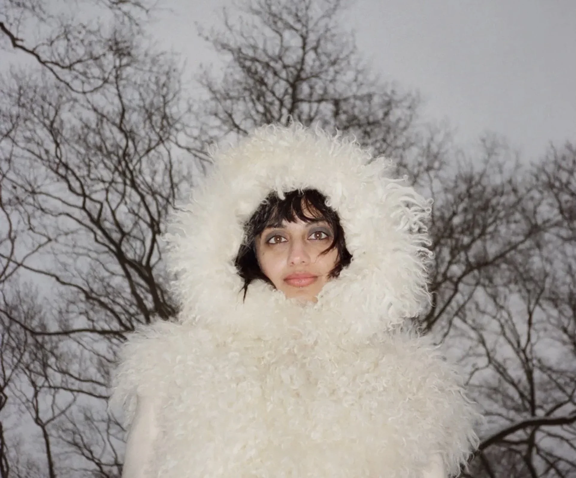 A woman with curly hair and brown eyes is standing outdoors in a snowy landscape with leafless trees in the background, wearing a white fluffy coat with a hood.
