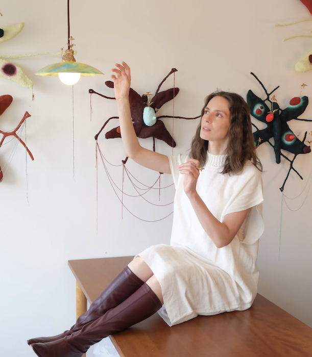 A woman with wavy brown hair sitting on a wooden table in front of a white wall decorated with hanging felt art insects. She is wearing a white dress and tall brown boots, and is reaching up with one hand.