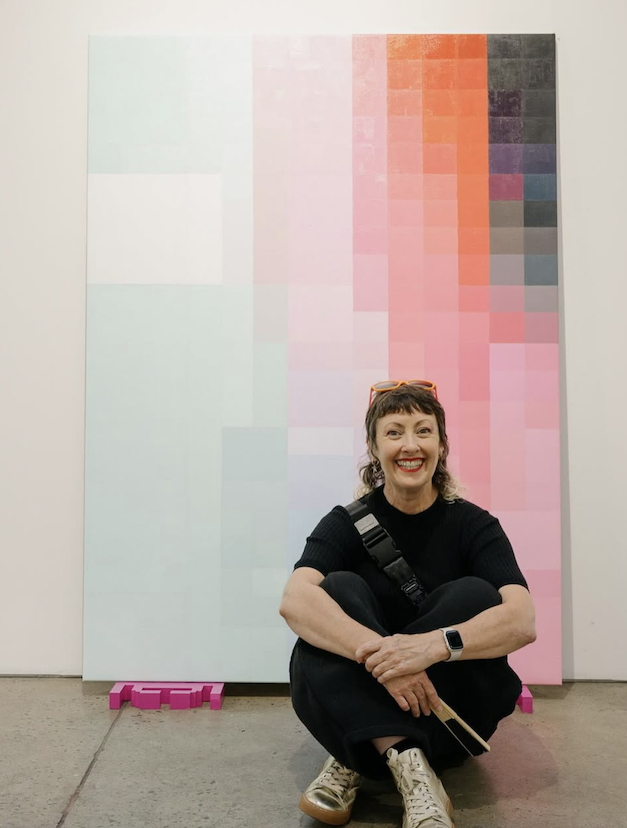 Woman sitting cross-legged in front of large color gradient artwork with pink, red, purple, gray, and black blocks, smiling at the camera.