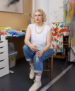 A woman with blonde curly hair is sitting on a wooden chair in an art studio. She is wearing a white sleeveless top, blue jeans, and white sneakers, with a pearl necklace. The background shows colorful fabrics, a whiteboard, and art supplies.