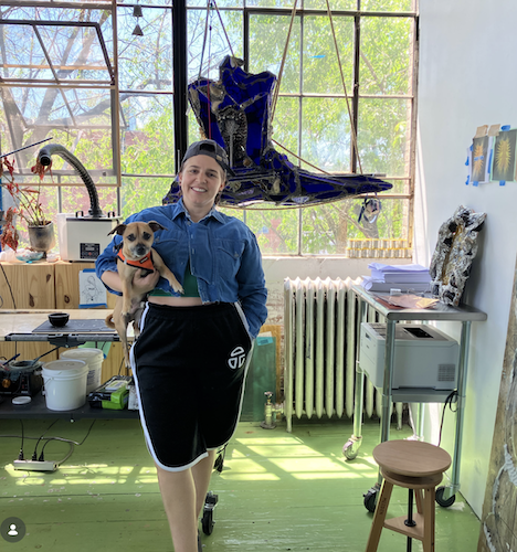 Woman smiling with a small dog in an art studio, colorful artwork and large windows in the background.