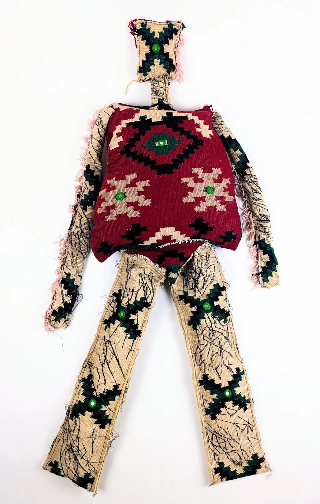 A fabric doll with a beard, wearing a red and black patterned shirt and beige pants decorated with black lines and green circular accents, against a plain white background.