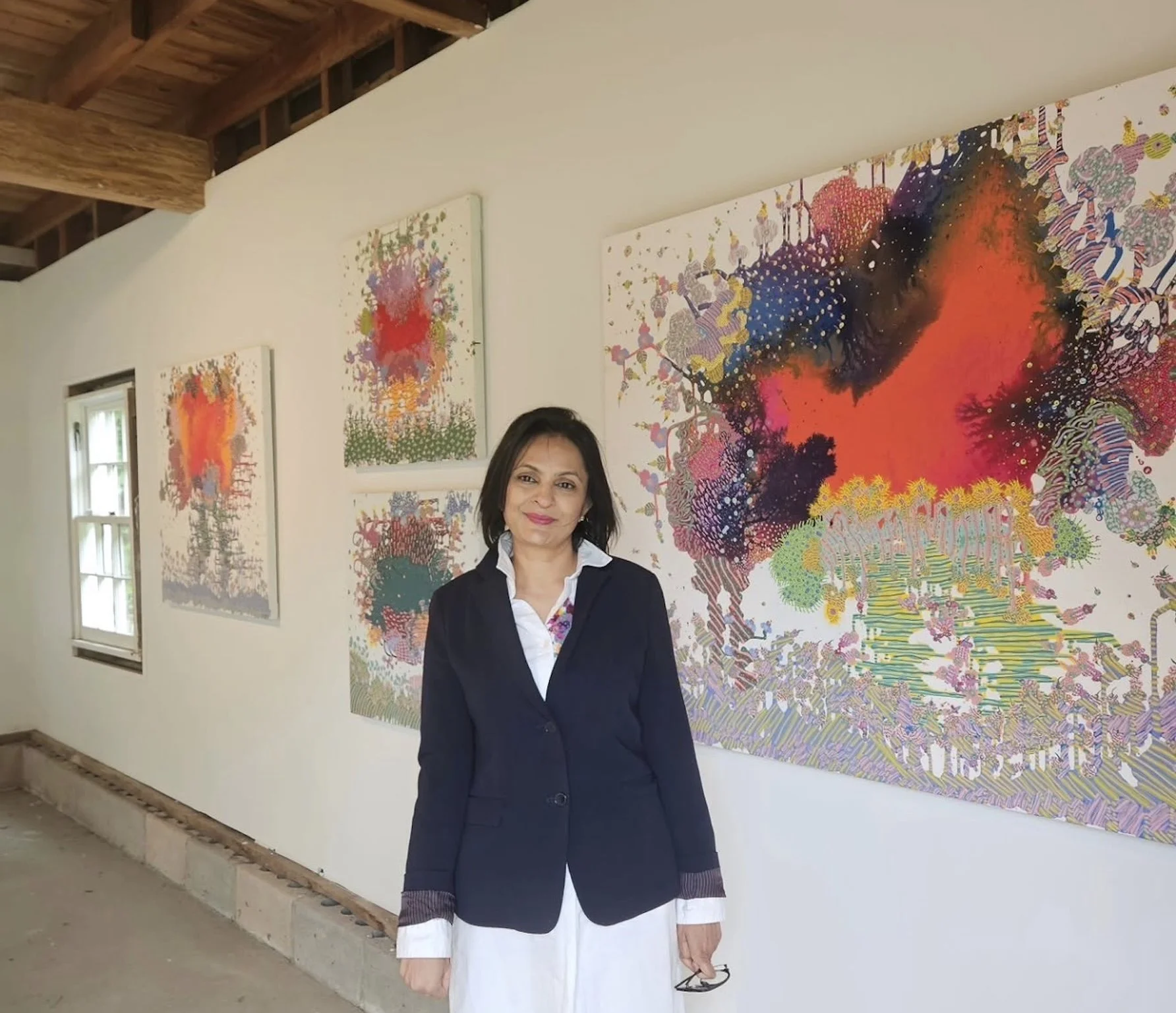 A woman standing in front of colorful abstract paintings on a white gallery wall. The woman is wearing a black blazer over a white dress and holding glasses in her right hand. The gallery has wooden ceiling beams and a window on the left side.