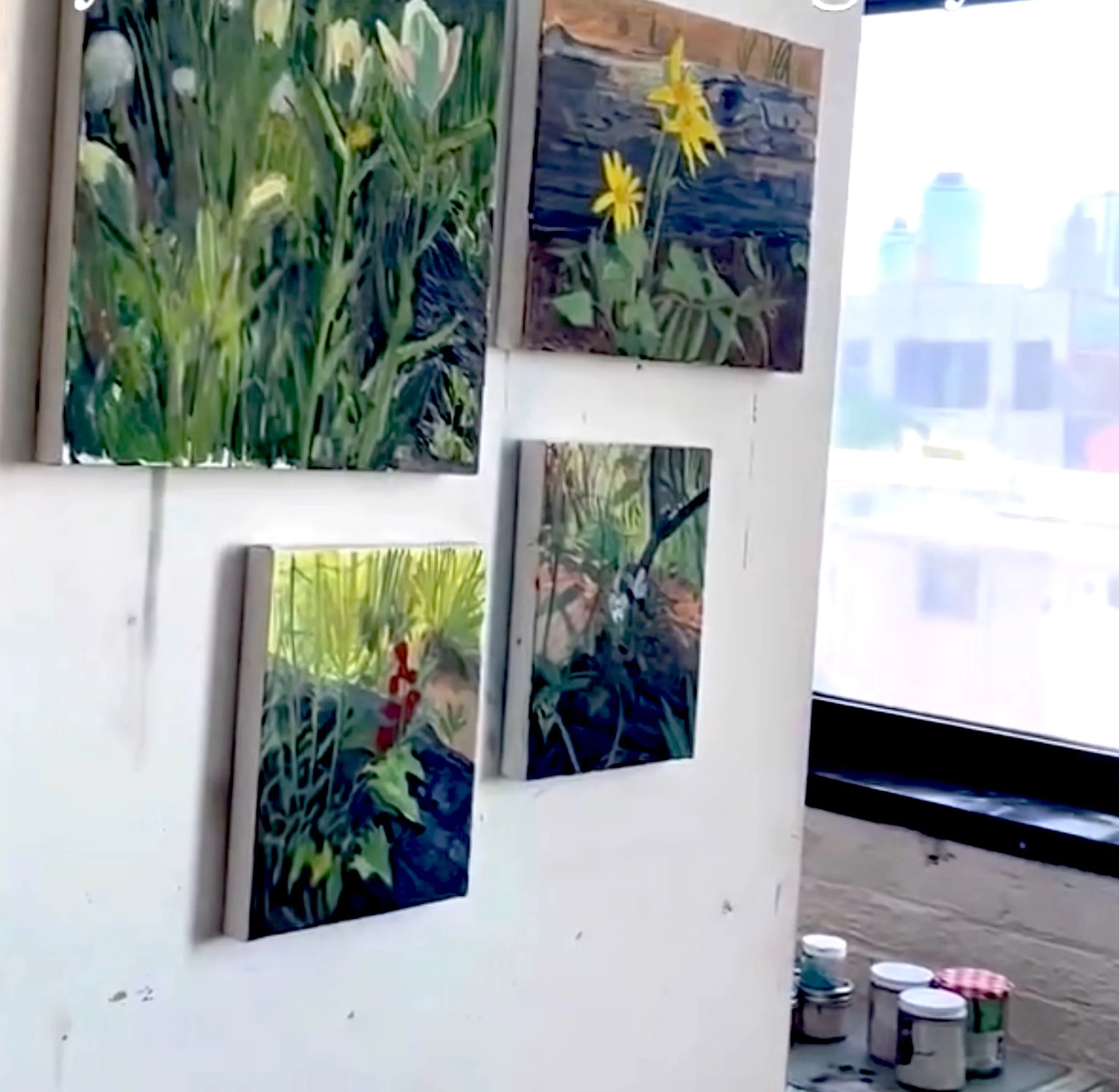 Multiple paintings of flowers and plants, displayed on a white wall in an art gallery, with some art supplies visible on a nearby table.