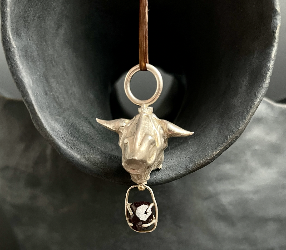 A dangling necklace featuring a silver cow skull pendant with a gemstone in the lower part, hanging from a leather cord inside a dark, curved ceramic vessel.