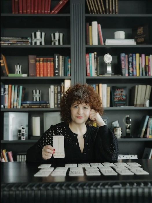 A woman with curly hair sitting at a table in front of a large bookshelf, holding a card, with more cards spread out on the table.